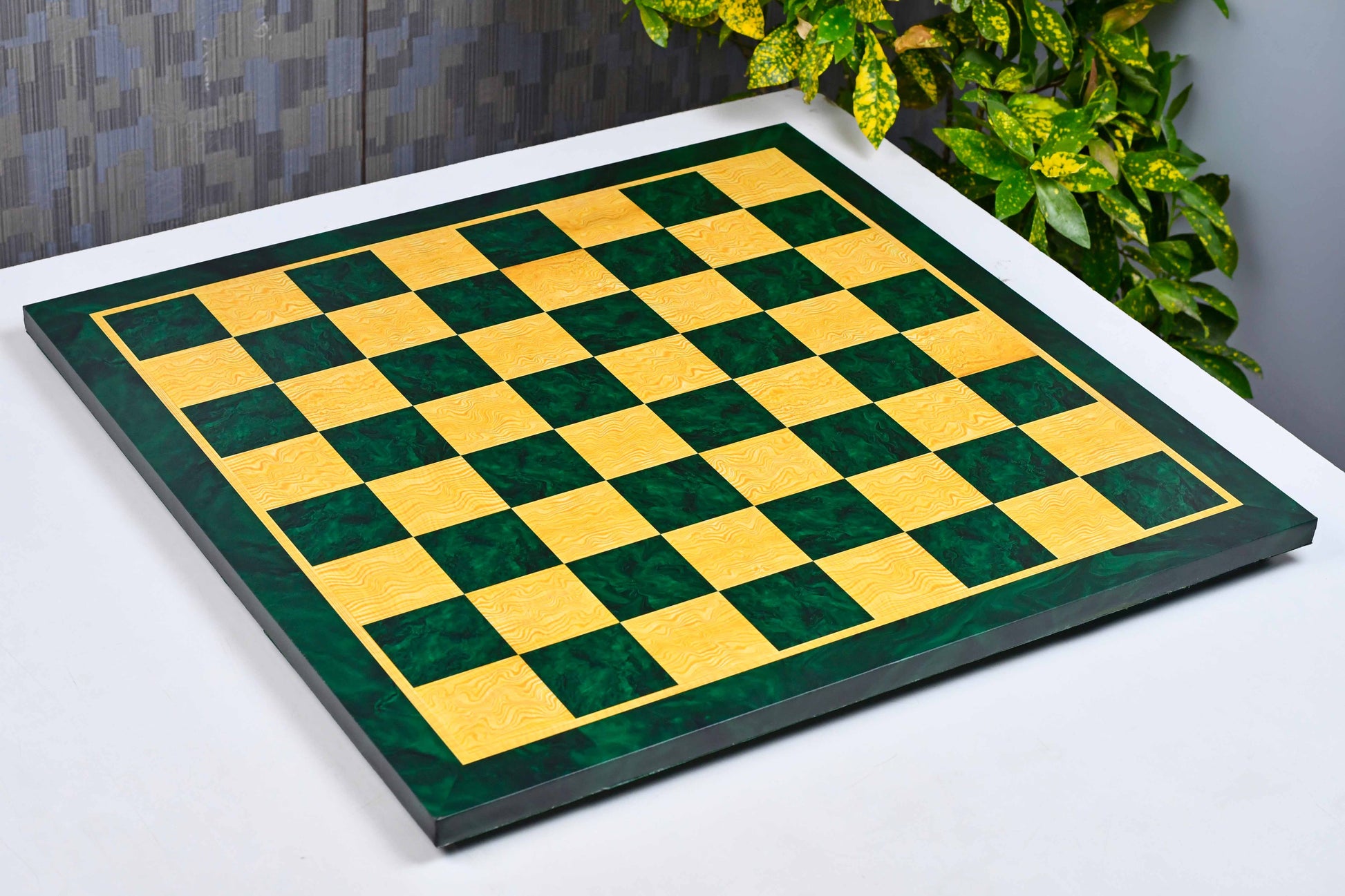 A square checkered board with alternating dark green and golden-yellow squares, bordered by a dark frame, shown resting on a white surface with a potted plant and textured wall in the background.
