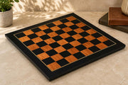 A square wooden chessboard with alternating black and warm brown checkers, framed by a black border with a thin gold line, set on a beige stone surface with a small plant and books in the background.