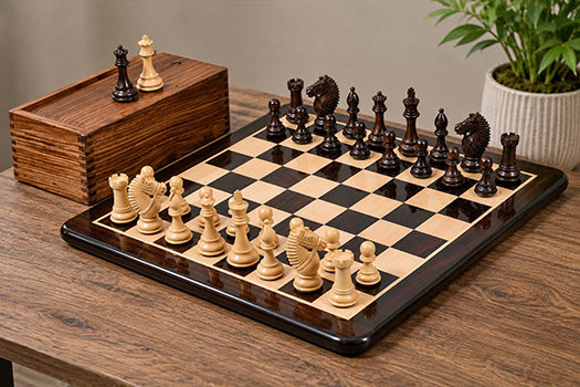 A wooden chessboard with black and cream chess pieces arranged on it, alongside a matching wooden storage box with two extra queens.